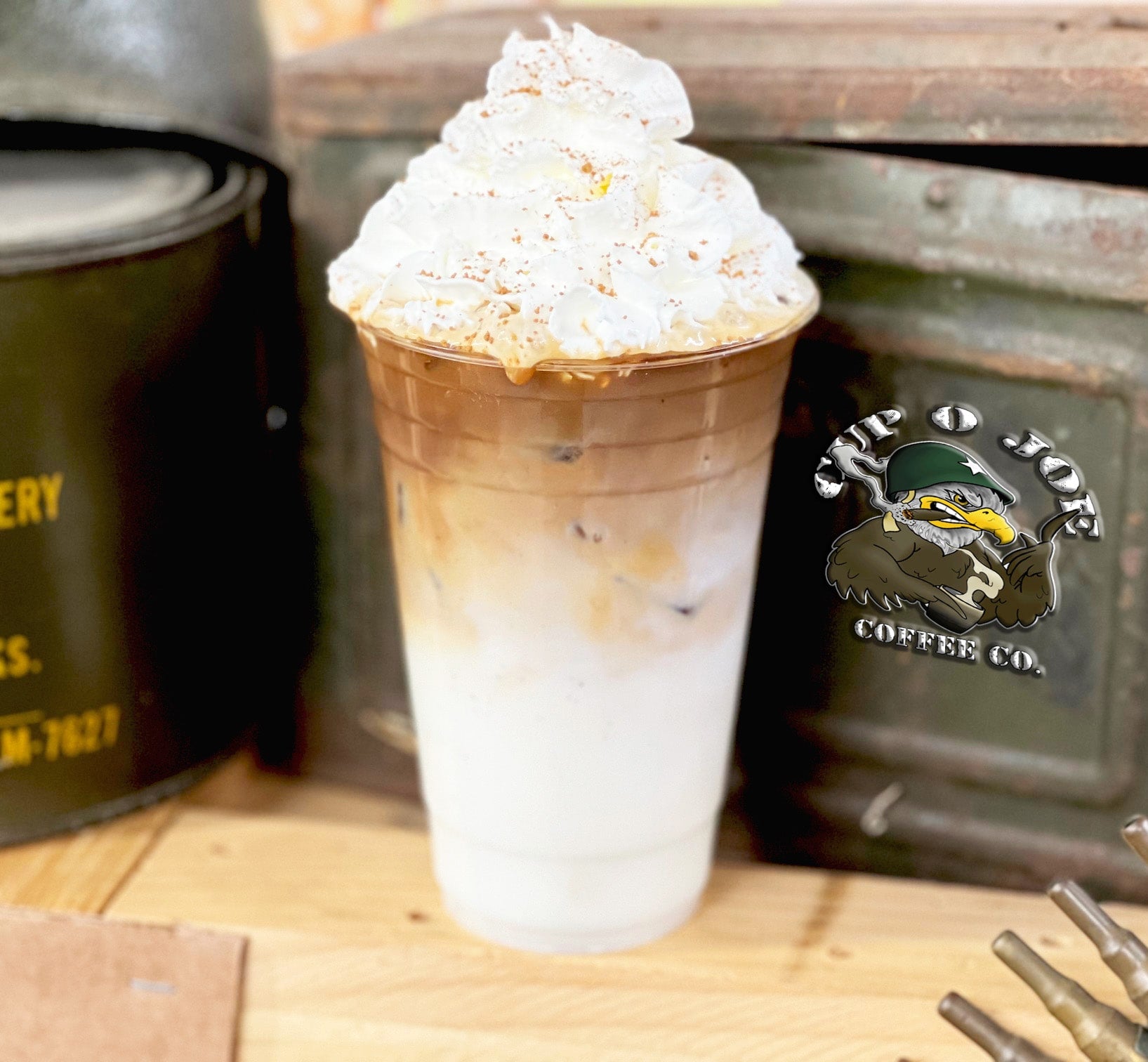 utter Pecan Macchiato served iced in a clear cup, topped with whipped cream and a sprinkle of cinnamon. The drink is placed on a rustic wooden table with military-inspired decor and the Cup O Joe Coffee Co. logo in the background.